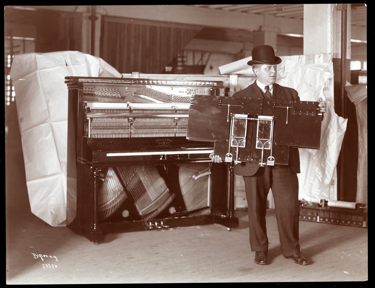 Ein Klavier von Ricca und Son Piano Co. mit einem Mann daneben, der einen Teil der Klavierwerke hochhält, 1907 von Byron Company