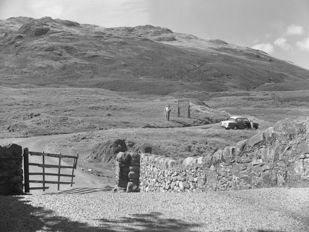 Blick auf die Straße über den Hard Knott Pass, zwei Personen stehen in der Nähe eines Straßenschilds und ein geparktes Auto auf dem Grasstreifen von Joseph Hardman