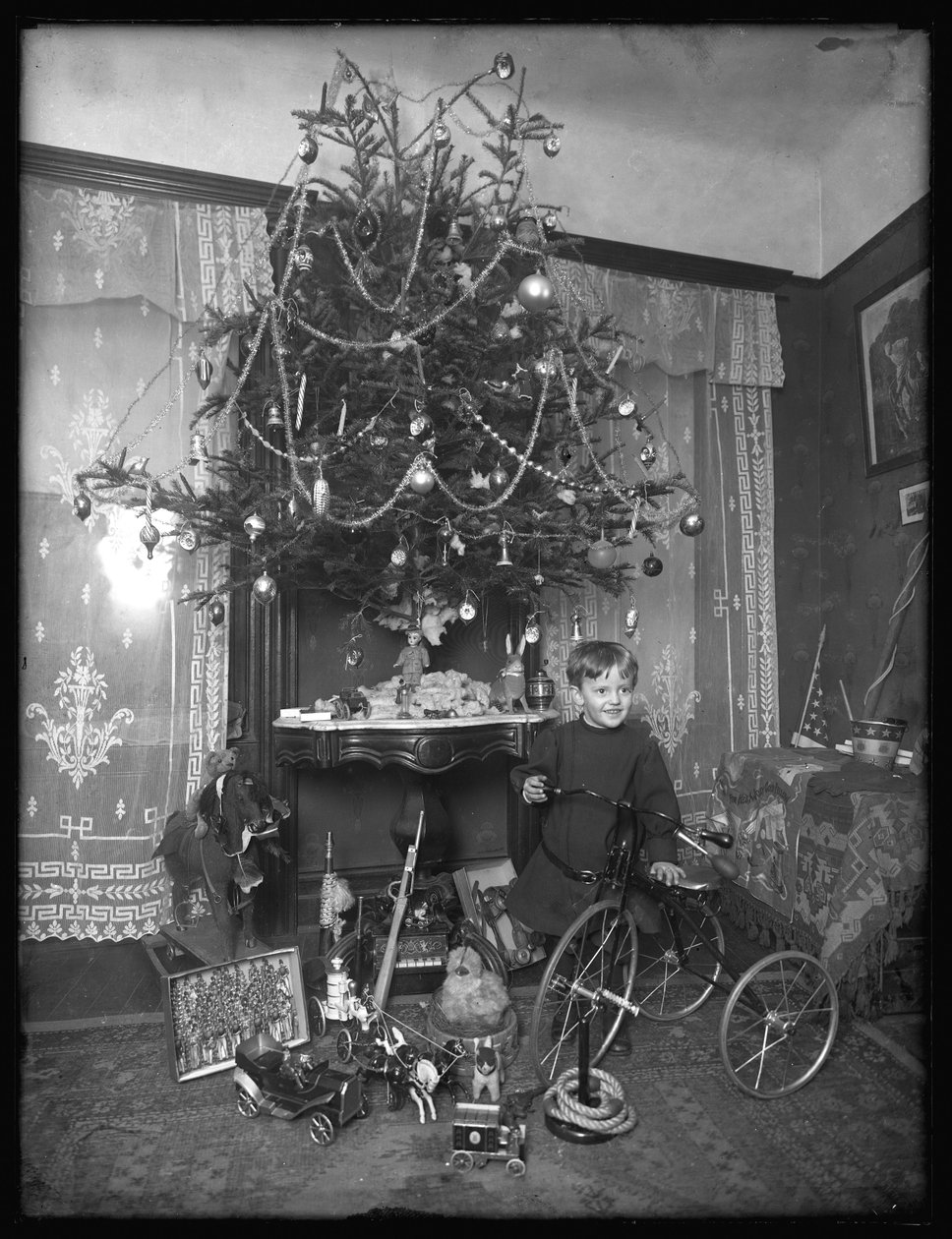 Seymour-Junge posiert mit Dreirad neben Weihnachtsbaum im Salon, Weihnachten 1912 von William Davis Hassler