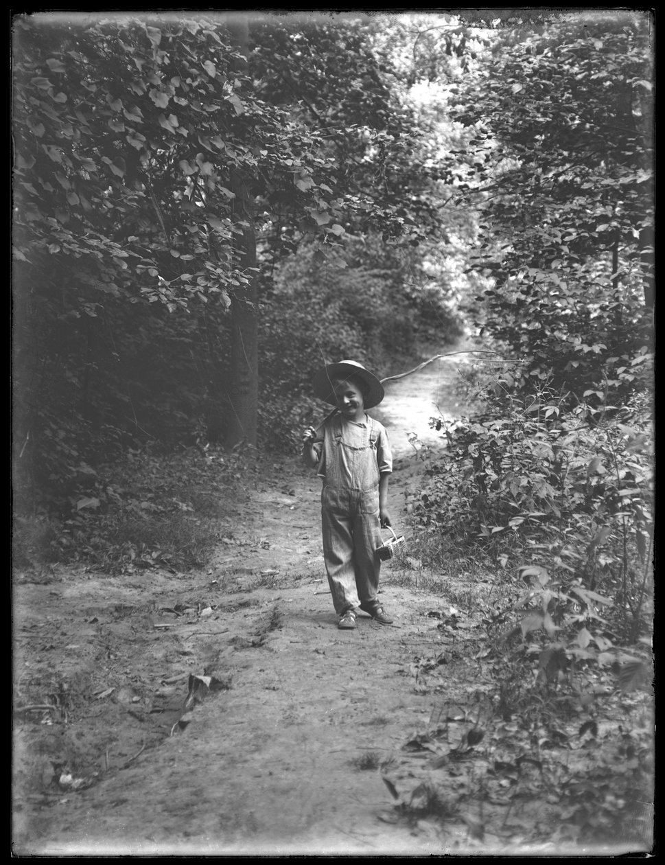 William Gray Hassler mit Strohhut und Latzhose, mit Angelrute und Korb, auf einem Waldweg gehend von William Davis Hassler