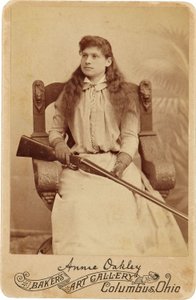 Annie Oakley: Hier sitzt die legendäre Scharfschützin eher schüchtern in einem massiven Renaissance-Stil Stuhl, ein Gewehr ruht auf ihrem Schoß. Ihre zarten weiblichen Züge und fließenden Haare täuschen über ihr robustes Outdoor-Image hinweg