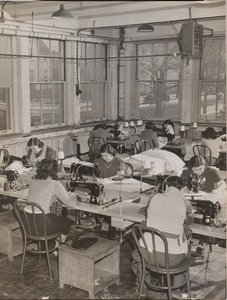 [Frauen sitzen an Schreibtischen und arbeiten an Nähmaschinen]
