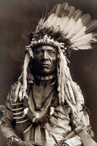 Wiesel Schwanz (Apohsuyis). Porträt des Indianerhäuptlings des Piegan-Stammes, trägt den berühmten Adlerfeder- und Wieselkopfschmuck, eine Wildledertunika, zwei Halsketten, eine aus Knochen und die andere aus Grizzlyklauen, und eine Tomahawk-Pfeife von Edward Sheriff Curtis