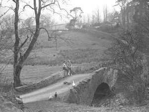 Zwei Mädchen sitzen auf einer Steinbrücke über den Fluss Cocker am Fuße des Scale Hill und beobachten Gänse in der Gasse bei Loweswater