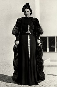 Modell im Abendkleid des italienischen Modehauses "Dragoni". Die Vorderseite des Artikels hat eine auffällige Paillettenverzierung in Form eines Schwertes. Ein Hut und ein Pelzmantel vervollständigen das Ensemble.