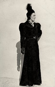 Modell präsentiert einen Artikel des italienischen Modehauses "Cicogna" bestehend aus einem Breitschwanzmantel, der an der Taille mit einem Gürtel mit juwelenbesetzter Schnalle zusammengehalten wird, ergänzt durch einen kleinen Federhut mit Schleier.