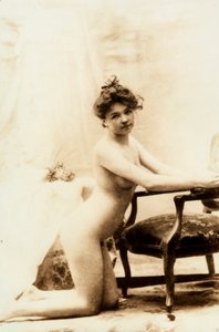 Nacktporträt einer jungen Frau, die vor einem kleinen Sessel kniet.