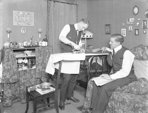 Unidentifizierter Mann, der die Verwendung eines elektrischen Bügeleisens demonstriert, beobachtet von einem anderen Mann, ca. 1913-14