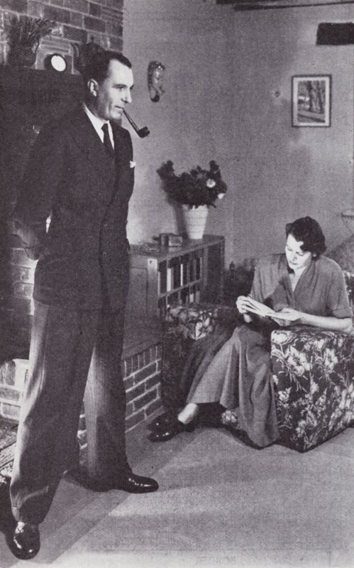 Frau liest, während der Mann sich am Feuer den Rücken wärmt, ca. 1950 von English Photographer