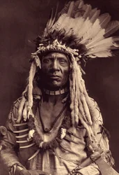 Wiesel-Schwanz (Apohsuyis). Dieser Piegan-Indianer trägt den berühmten Kriegsschmuck aus Adlerfedern und Wieselfell, eine Wildleder-Tunika, zwei Halsketten, eine aus Knochen und die andere aus Grizzly-Klauen, und eine Tomahawk-Pfeife. Foto aus Band 6 der E