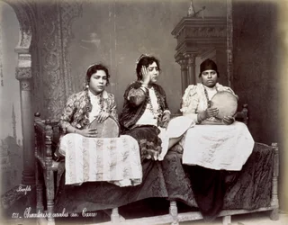 Porträt von drei jungen arabischen Sängerinnen in ethnischer Kleidung mit langen Halsketten. Sie posieren im Studio, sitzend. Zwei von ihnen halten ein Tamburin