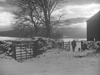 Mann und Hund gehen durch ein Tor zur Kamera im Schnee mit Thirlmere dahinter