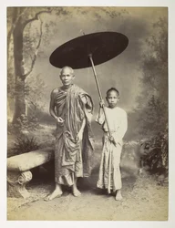 Burmesischer Mönch und Begleiter, Ganzkörper-Studio-Porträt, vor einer gemalten Kulisse einer Waldszene, der Mönch wird von einem jungen chinesischen Diener begleitet, der einen Regenschirm über ihn hält