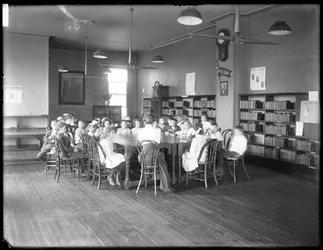 Kinder hören einer Frau im Lesesaal einer nicht identifizierten Zweigstelle der Queens Borough Public Library zu, um 1910