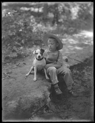 William Gray Hassler mit Strohhut und Latzhose, sitzend im Wald mit einem kleinen Hund (Bessie), 20. August 1911
